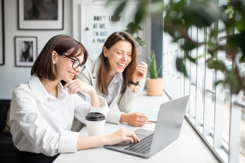 Boost Productivity: Two happy and smiling remote female workers looking at a laptop in a dedicated workspace